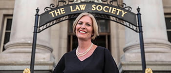 Christina Blacklaws, a white woman with chin-length straight blonde hair, stands smiling widely. She wears a black dress and a pearl necklace.