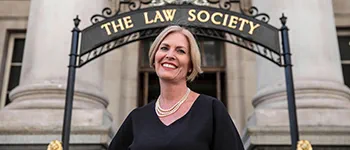 Christina Blacklaws, a white woman with chin-length straight blonde hair, stands smiling widely. She wears a black dress and a pearl necklace.