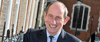 Michael Napier, a white man with short dark grey hair, stands smiling widely. He wears a navy suit, light blue shirt and dark blue tie.
