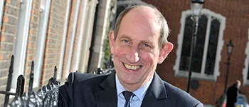 Michael Napier, a white man with short dark grey hair, stands smiling widely. He wears a navy suit, light blue shirt and dark blue tie.