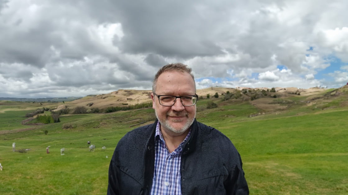 Brett Dixon is a white man with short dark grey hair, glasses and a short grey beard. He is smiling and standing in a windswept rural landscape, wearing a navy jacket and a blue and white checked shirt.