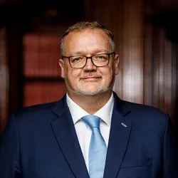 Brett Dixon stands smiling in the Law Society library in London. Brett is a white man with short dark grey hair, a cropped grey beard and glasses. He wears a navy suit, white shirt and light blue tie.