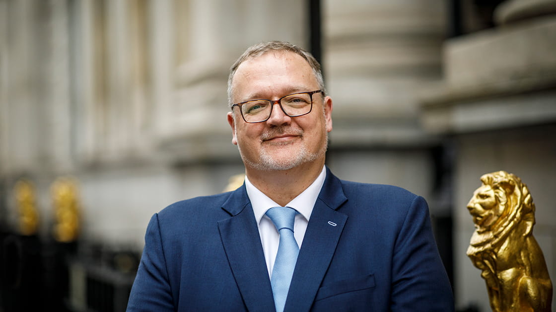 Brett Dixon stands smiling in front of the Law Society on Chancery Lane. Brett is a white man with closely cropped grey hair, a short grey beard and glasses. He is wearing a navy suit, white shirt and pale blue tie.