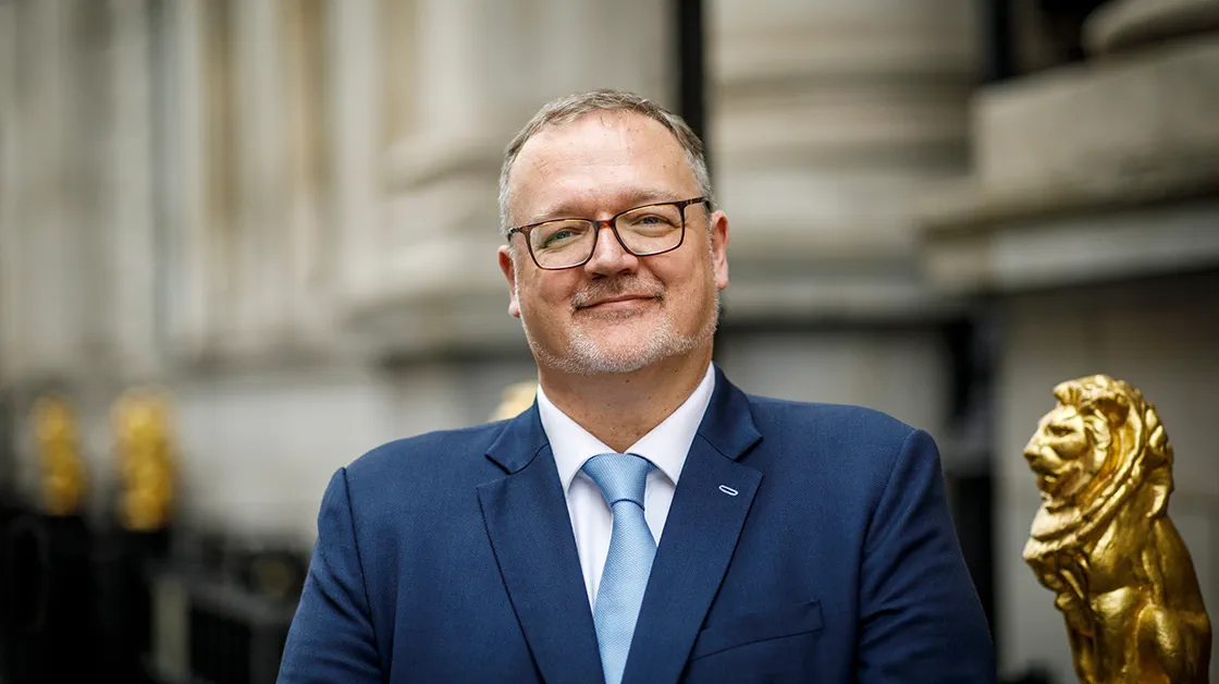 Brett Dixon stands smiling in front of the Law Society on Chancery Lane. Brett is a white man with closely cropped grey hair, a short grey beard and glasses. He is wearing a navy suit, white shirt and pale blue tie.