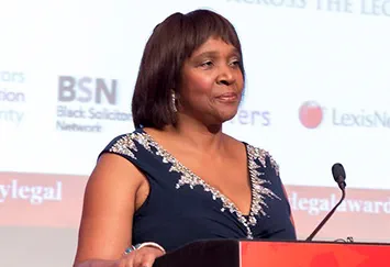 Cordella Bart-Stewart OBE is a black woman with shoulder-length straight dark brown hair. She stands at a podium speaking at a Black Solicitors Network event.