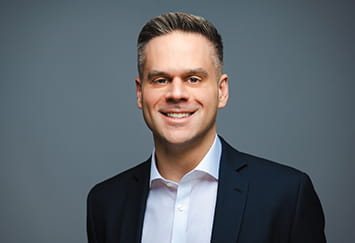 Matt Aleksic is a white man with short, straight brown hair. He is smiling widely and wears a black t-shirt.