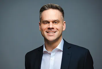 Matt Aleksic is a white man with short, straight brown hair. He is smiling widely and wears a black t-shirt.