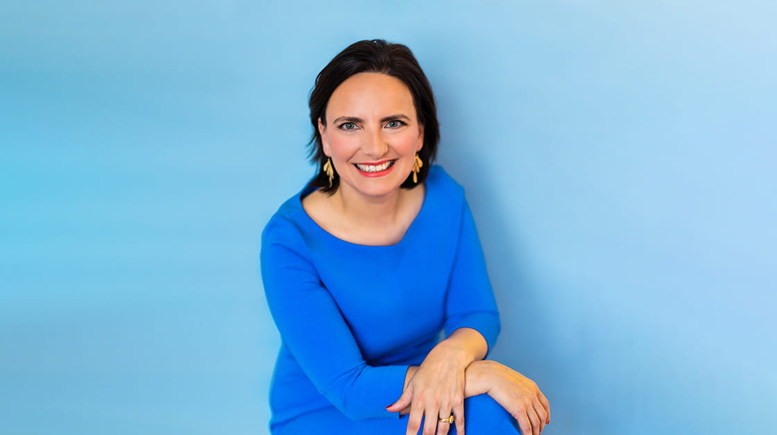 Dana Denis Smith is a white woman with dark, shoulder-length straight hair. She sits smiling and wearing an electric blue dress with gold jewellery.