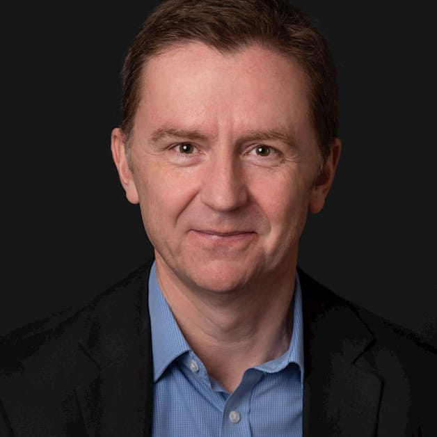 Ian Jeffery is a white man with short brown hair. He is smiling and wears a blue shirt and black jacket against a black background.