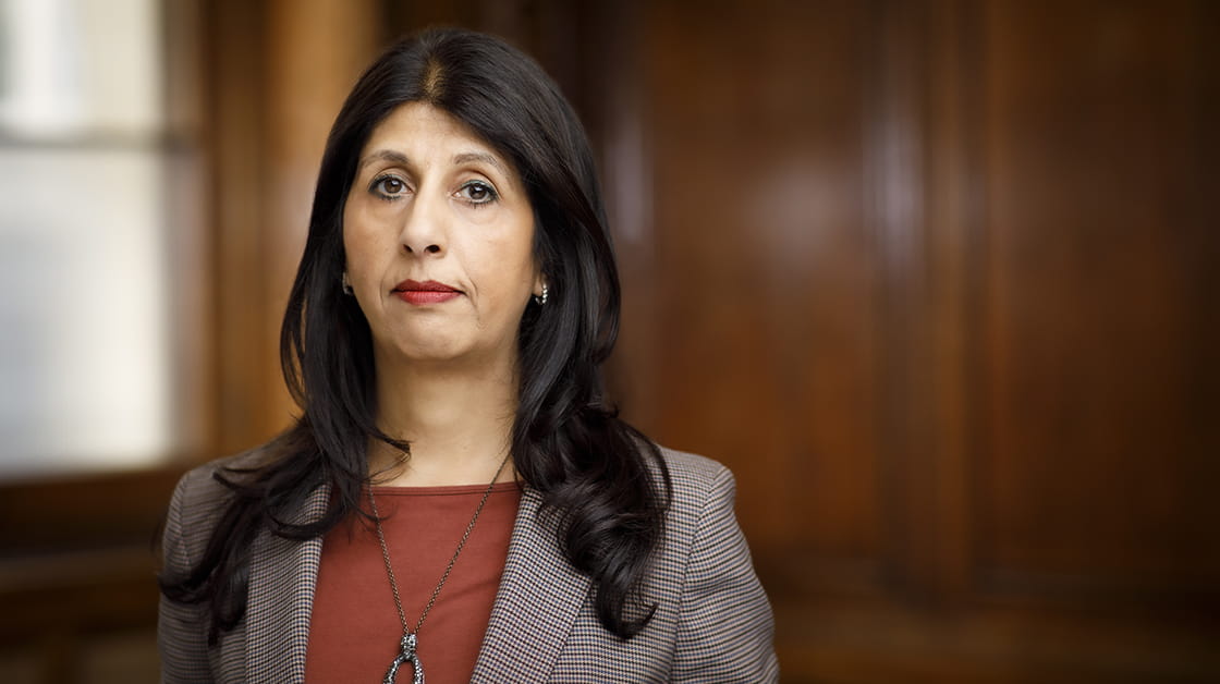 Lubna Shuja is a woman of South Asian heritage with long, wavy, dark brown hair. She looks serious and wears a grey suit and burnt orange shirt.