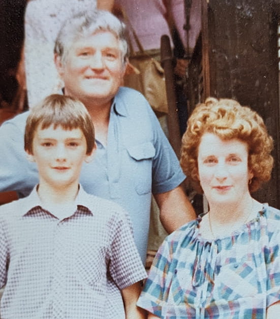Mark Evans as a young boy smiling with his dad and mum. Mark is white and has short dark brown hair and wears a short sleeve check shirt. His dad is a white man with grey hair and wears a blue short sleeve shirt. His mum is a white lady with a short brown perm, she wears a tartan summer dress. 