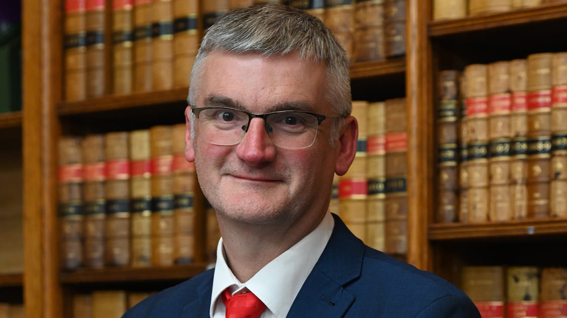 Mark Evans stands in the Law Society library. He is a white man with short grey hair, smiling and wearing a navy suit, white shirt and red tie.