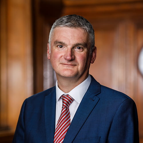 Mark Evans stands in a panelled room at the Law Society in London. Mark is a white man with short dark grey hair. He wears a navy suit, white shirt and red, white and silver striped tie.