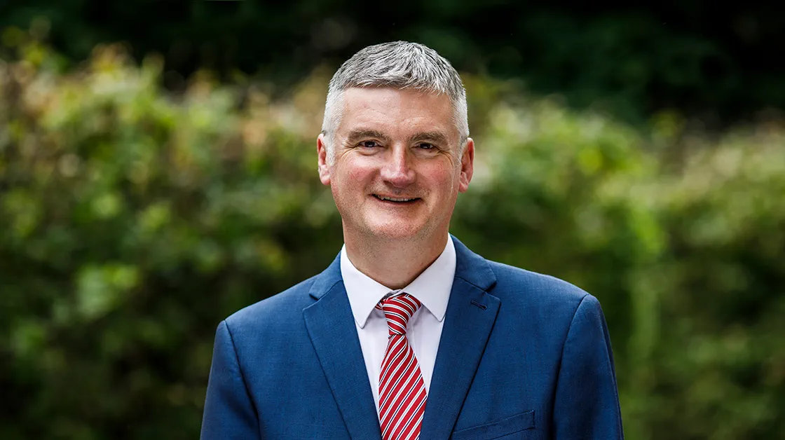 Mark Evans stands outside, smiling widely. Mark is a white man with short grey hair. He wears a navy suit, white shirt and red, white and silver striped tie.