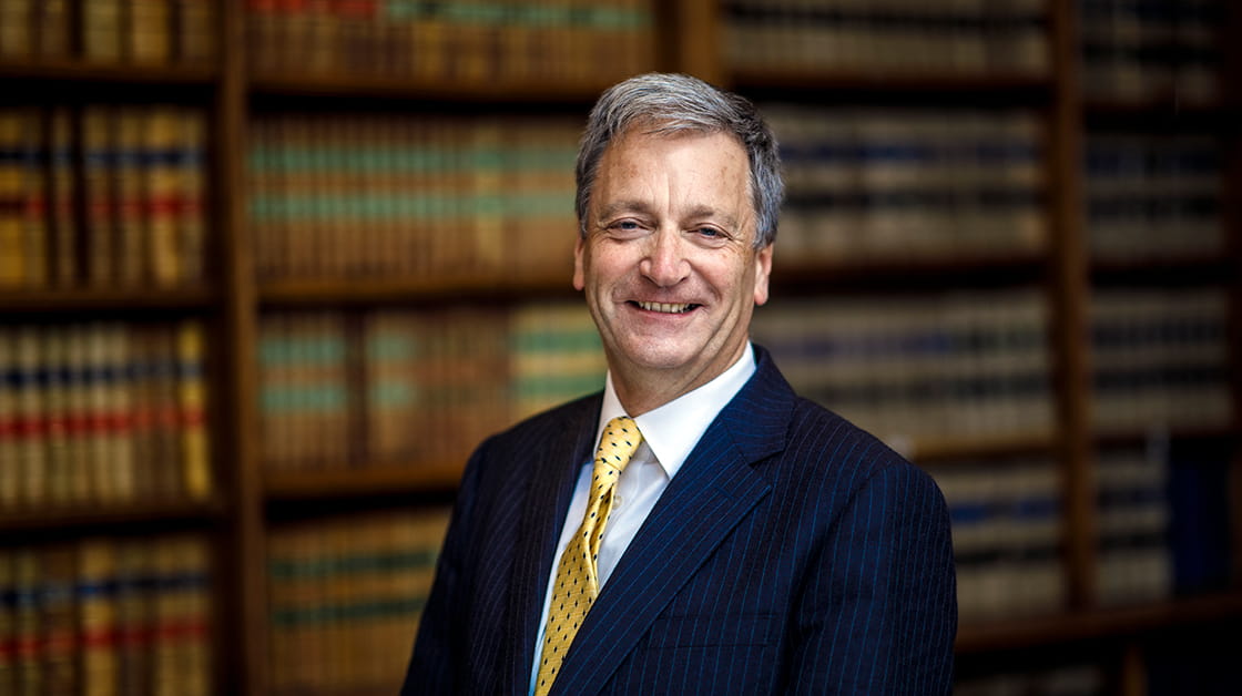 Richard Atkinson stands smiling in the Law Society library. Richard is a white man with short dark grey hair. He wears a navy pinstriped suit, white shirt and gold polka dot tie.