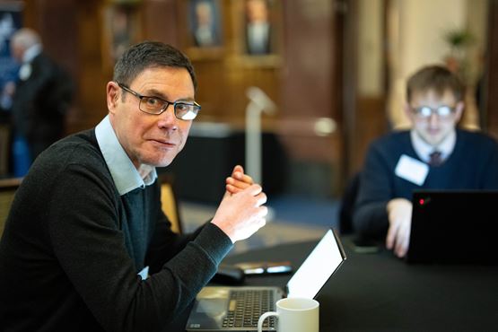 Man sitting at a laptop and wearing glasses, a blue shirt and a black jumper