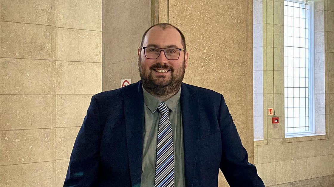 Gary is a white man, he's wearing glasses, a navy blue jacket, green shirt and blue stripy tie. He is standing and smiling in a stone building, with a window behind him. 