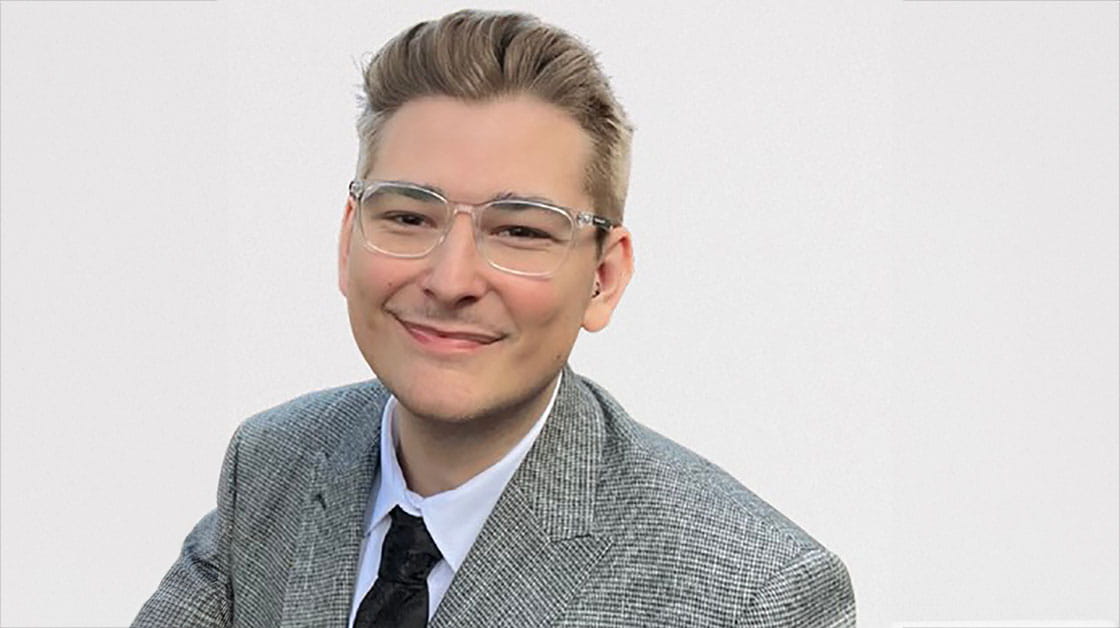 Josh Ackroyd is a white man with blonde hair and glasses. He is wearing a grey suit jacket, white shirt and black tie. He is smiling with his mouth closed in front of a white background.