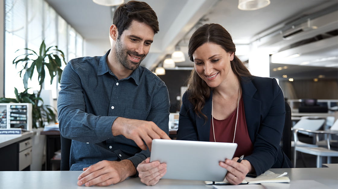 A man with dark hair and blue shirt sits next to a woman with long brown hair and a purple blouse, in an office. They are both looking at a tablet and smiling.