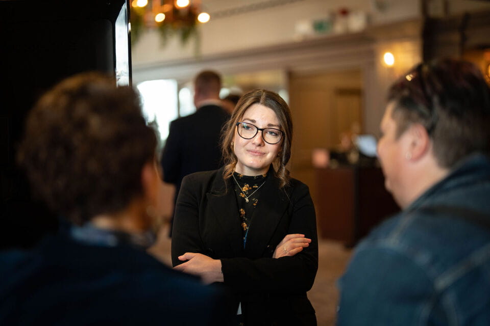 A white woman with brown hair and glasses is wearing a black jacket. She has her arms crossed and is talking to men.