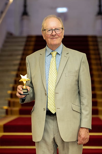 Clive Sutton, Legal Hero 2023. Clive is a white man with short grey hair and glasses. He is smiling and stands on the grand staircase at 113 Chancery Lane, London holding his award. He wears a light linen suit, blue shirt and gold tie with a navy polka dot pattern.