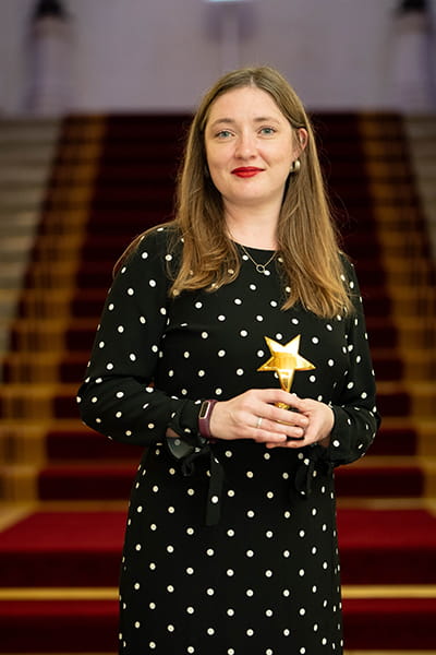 Emily McFadden, Legal Hero 2023. Emily is a white woman with long, straight brown hair. She is smiling and stands on the grand staircase at 113 Chancery Lane, London holding her award. She wears a long, black dress with white polka dots.