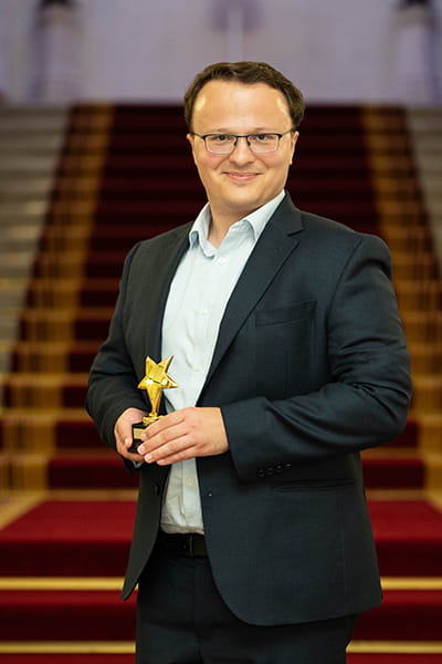 Martin Whitehorn, Legal Hero 2023. Martin is a white man with short, wavy brown hair and glasses. He is smiling and stands on the grand staircase at 113 Chancery Lane, London holding his award. He wears a black suit and open-collar white shirt.