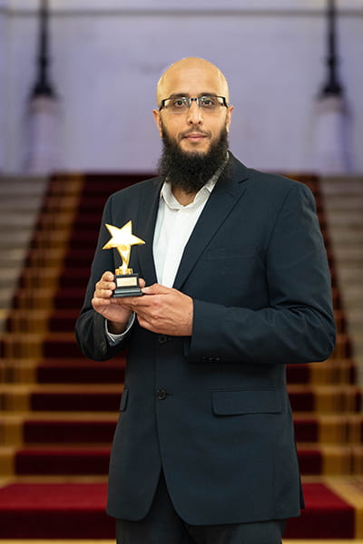 Qaisar Sheikh, Legal Hero 2023. Qaisar is a bald man with a long black beard and glasses. He is smiling and stands on the grand staircase at 113 Chancery Lane, London holding his award. He wears a black suit and open-collar white shirt.