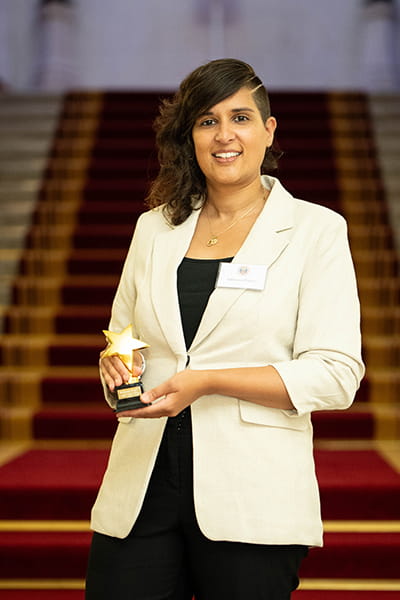 Sabeena Pirooz, Legal Hero 2023. Sabeena is a woman of South Asian heritage with long, curly, dark brown hair and a buzzcut. She is smiling and stands on the grand staircase at 113 Chancery Lane, London holding her award. She wears a cream blazer over a black shirt and suit trousers.