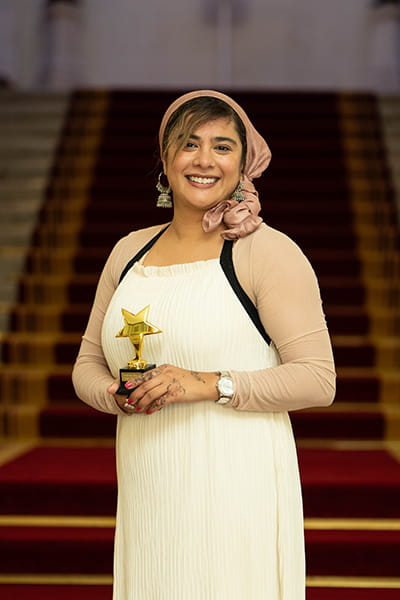 Shabina Begum, Legal Hero 2023. Shabina is a woman of South Asian heritage with wavy brown hair, which she wears under a dusty pink headscarf. She is smiling and stands on the grand staircase at 113 Chancery Lane, London holding her award. She wears a long white dress with black straps and long beige sleeves.