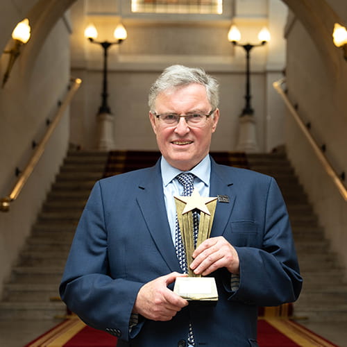 Duncan McNair, Legal Hero 2024. Duncan is a white man with short light grey hair wearing glasses and a navy suit, light blue shirt and navy tie emblazoned with elephants. He stands on the Law Society steps holding his award, a golden starburst.
