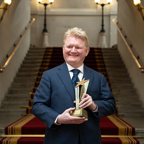 Kevin McManamon, Legal Hero 2024. Kevin is a white man with short light auburn hair wearing a navy suit, white shirt and navy tie. He stands on the Law Society steps holding his award, a golden starburst.