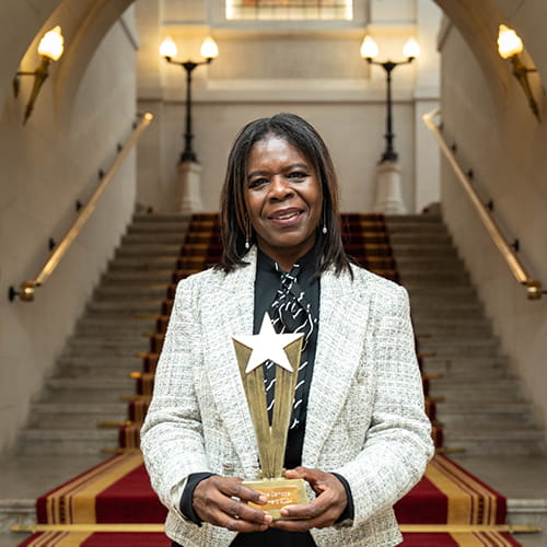 Pauline Campbell, Legal Hero 2024. Pauline is a black woman with shoulder-length straight brown hair wearing a white tweed suit, black shirt and black-and-white striped cravat. She stands on the Law Society steps holding her award, a golden starburst.