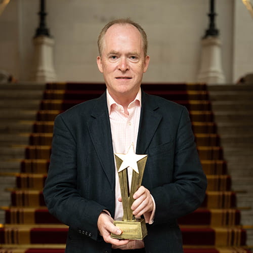 Stephen Kinsella, Legal Hero 2024. Stephen is a white man with closely cropped brown hair, wearing a black suit and peach-and-white checked open collar shirt. He stands on the Law Society stairs holding his award, a golden sunburst.