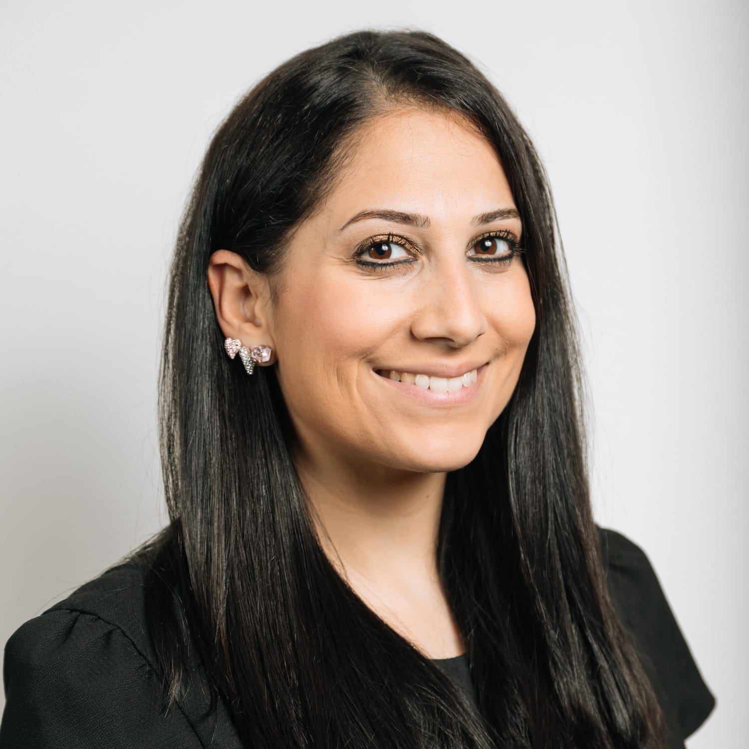 Alexia Eliades is a tanned woman with long, straight black hair. She is smiling widely and wears a black cap-sleeve dress and a variety of silver earings.