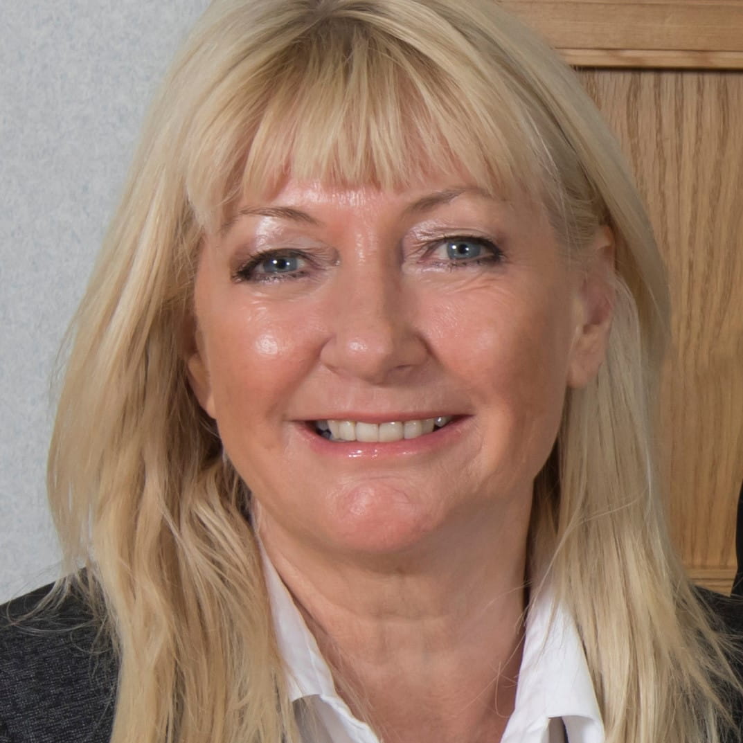 Hilary Meredith-Beckham is a white woman with long, straight blonde hair. She is smiling widely and wears a dark grey suit over a white open-collar shirt.