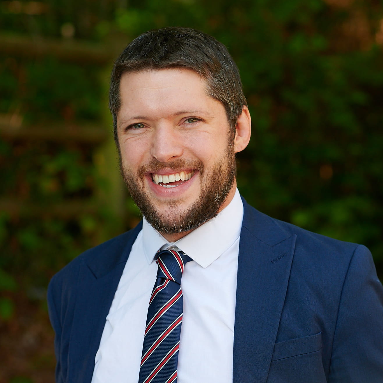 Jonathan Whettingsteel is a white man with cropped dark brown hair and a short brown beard. He is smiling widely and wears a navy suit, white shirt and a dark blue, white and red striped tie.