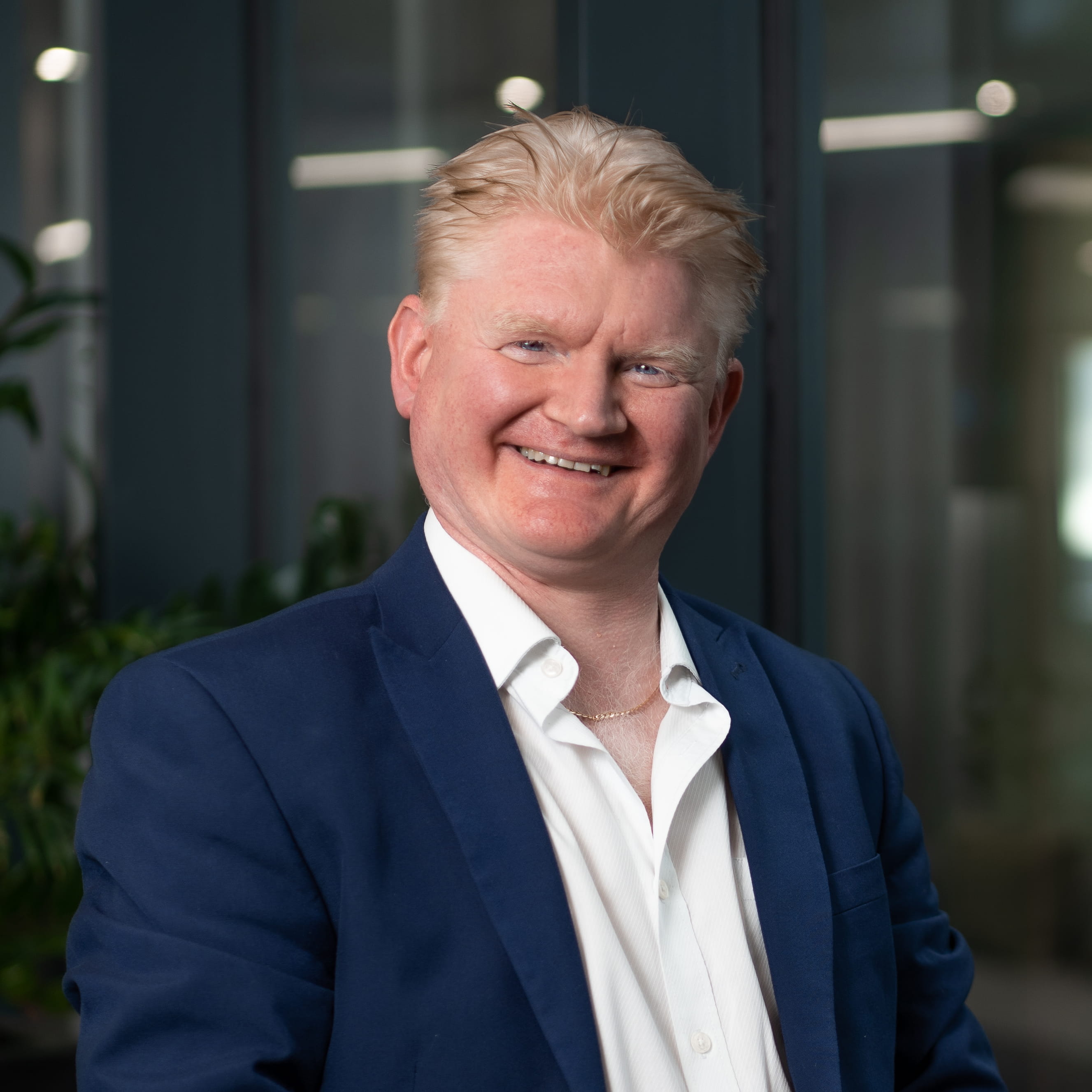 Kevin McManamon is a white man with short, wavy auburn hair tinged with grey. He is smiling widely and wears a navy suit, open collar white shirt and a gold chain.