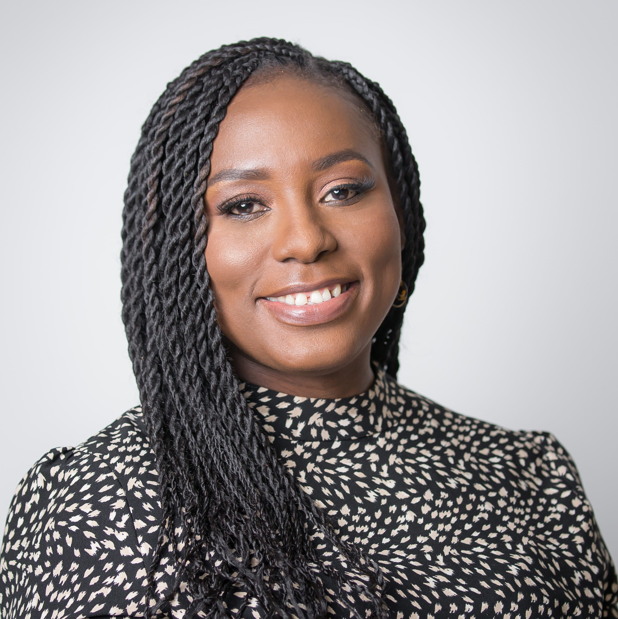 Kishma Bolaji is a black woman with long braided black hair. She is smiling widely and wearing a black-and-white patterned blouse.