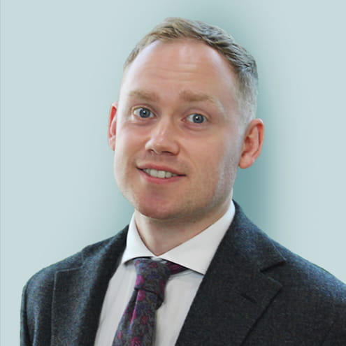 Michael Walker is a white man with cropped dark blonde hair. He is smiling and wears a dark grey suit, white shirt and grey and purple patterned tie.