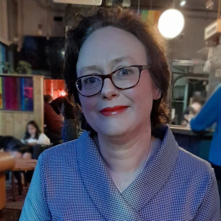 Michelle Waite is a white woman with short, wavy dark brown hair. She is smiling and wears a houndstooth jacket with dark-framed glasses and red lipstick.