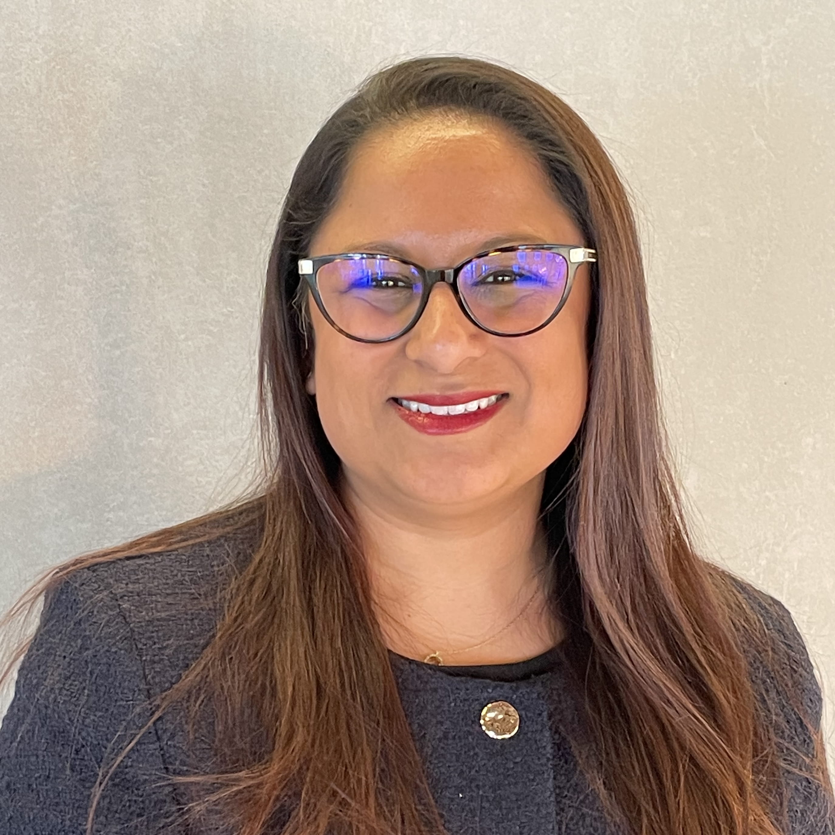 Reena Parmar is a woman of South Asian heritage with long, straight dark brown hair. She is smiling widely and wears a navy jacket, dark-framed glasses and a gold chain necklace.