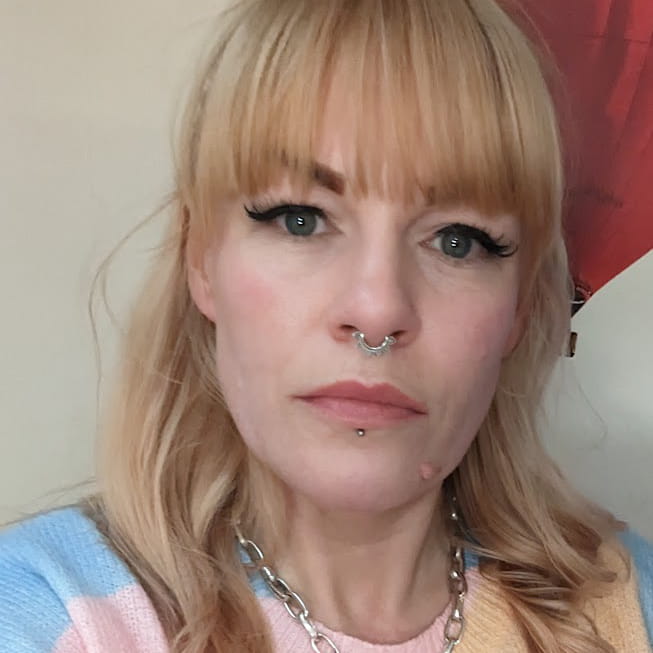 Siobhan Taylor-Ward is a white woman with long, wavy blonde hair. She wears a pastel blue and pink patterned shirt, a silver septum piercing, a silver lip piercing and a silver chain necklace
