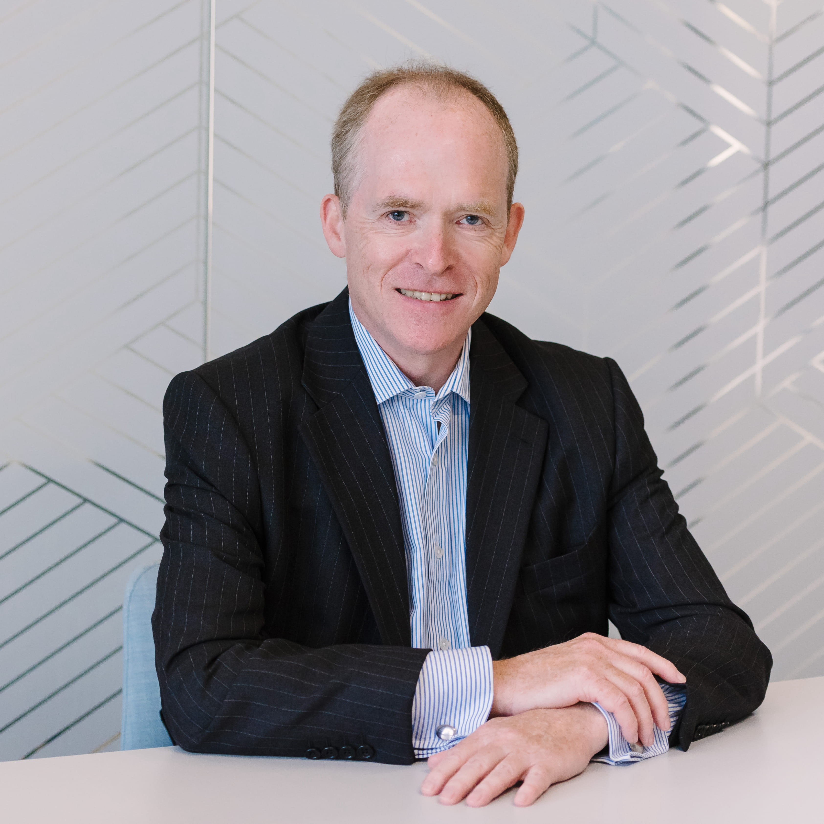 Stephen Kinsella is a white man with closely cropped dark hair. He is smiling widely and wears a black pinstriped suit and a blue-and-white striped open collar shirt.