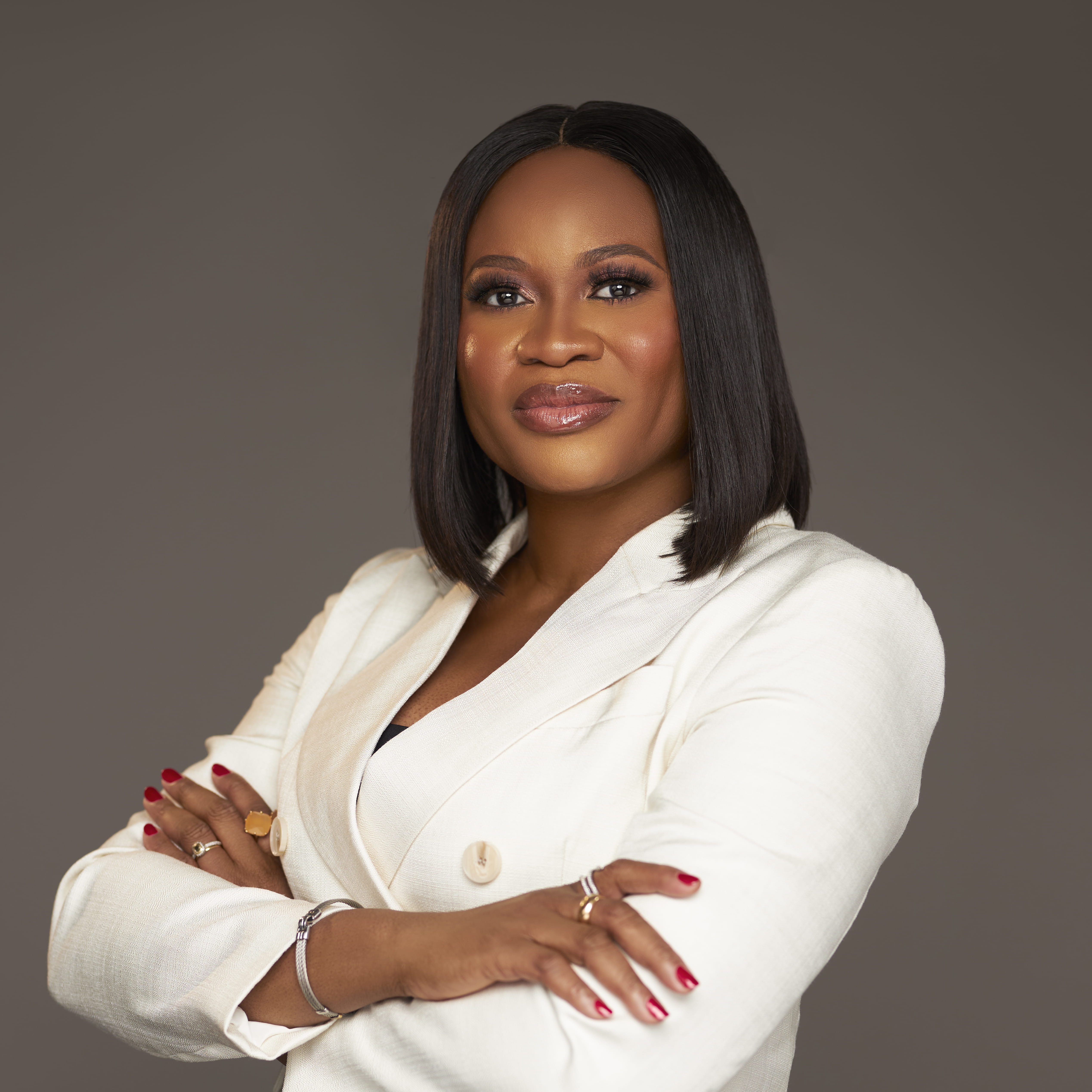 Tamara Egbedi is a black woman with short, straight black hair. She is smiling and stands with her arms crossed, wearing a white suit with a silver bangle and gold and silver rings.