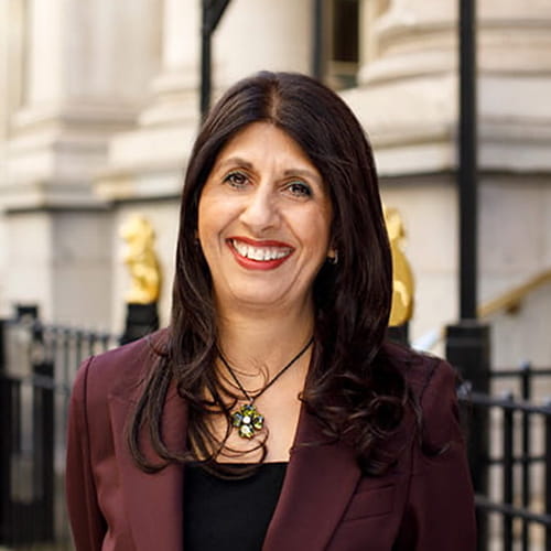 Lubna Shuja smiles widely, wearing a burgundy suit and a green necklace.