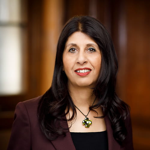 Lubna Shuja smiles, wearing a burgundy suit and a green necklace.