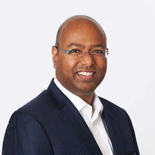 Dr Sam De Silva smiles, wearing a navy pinstriped suit and glasses.