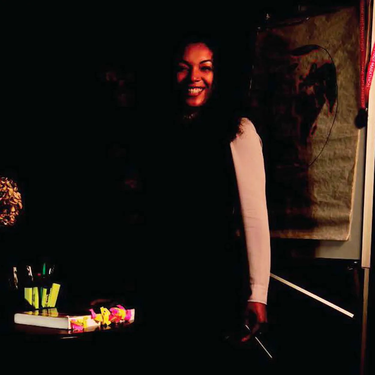 Adele Edwin-Lamerton is a Black woman with long, dark, wavy hair. She is smiling and wears a long-sleeved white blouse and black dress. She stands holding a pen in front of a desk with a heavily tabbed textbook.