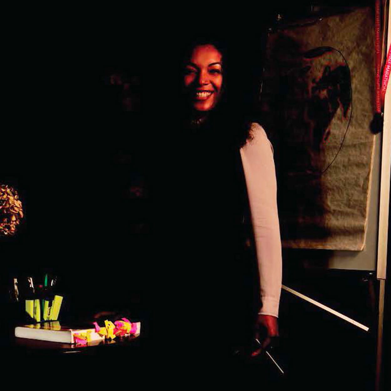 Adele Edwin-Lamerton is a Black woman with long, dark, wavy hair. She is smiling and wears a long-sleeved white blouse and black dress. She stands holding a pen in front of a desk with a heavily tabbed textbook.